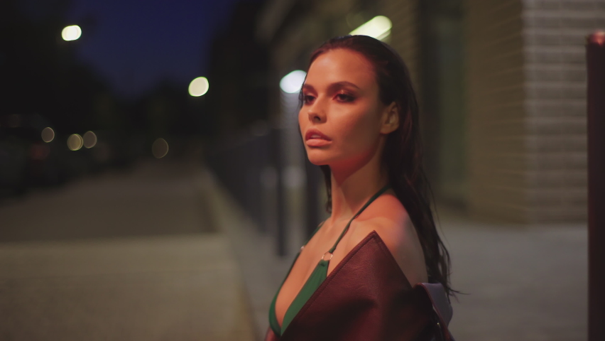 Close-up portrait young fashion woman sitting on the sidewalk and relaxing urban background of modern night city. Beautiful party girl in brown jacket and green bra woman looking around 4K