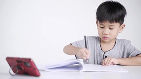 Asian boy about 5 years old writing while watching a mobile phone as studying online due to Covid 19 pandemic.
