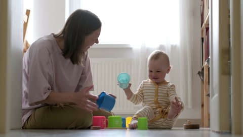 Little baby girl and mommy play with colorful toys at home sitting on floor. Mother and daughter laughing having fun together.