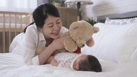 joyful asian new mom is holding and waving the stuffed toy’s hand while she is leaning close to her baby and interacting with it on the bed at home.