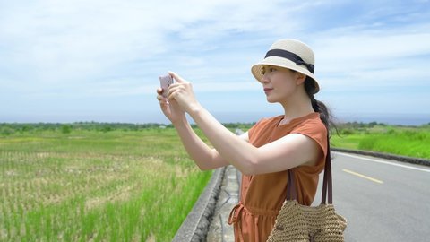 pretty Taiwanese lady wearing a hat is using her phone taking photos of the open green field with joy in the countryside of King Kong Avenue on a clear day.