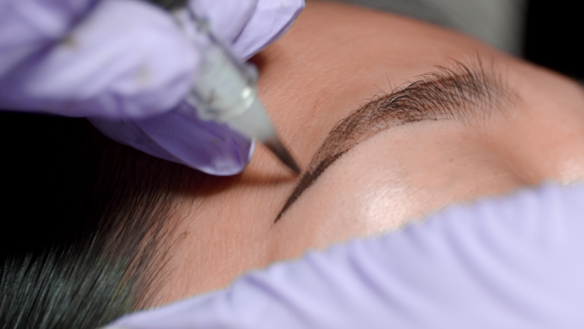Close up microblading eyebrow tattoo. Beautician in gloves makes permanent makeup correction of the shape of the eyebrows to a young women. Eyebrow tattoo in a beauty salon. | Shutterstock HD Video #1078826354