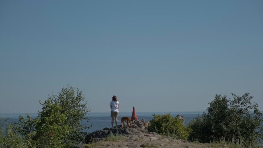 A young woman travels along the coast of the sea with a red dog of the Pharaoh breed. Summer sunny day, blue sky and stones.