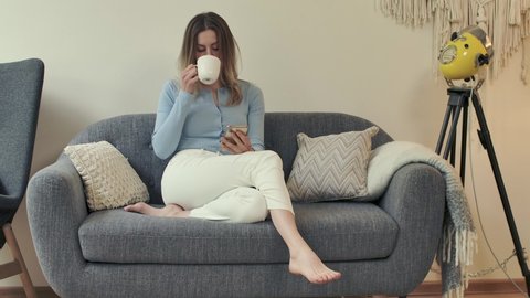 Cute cheeky female person sits on cozy sofa at her apartment with smartphone and coffee cup in hands. Bare feet woman scrolls and browses, sips drink. Charming girl texting while lying on comfy couch.