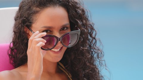 Headshot portrait of happy cheerful curly Hispanic girl student takes off sunglasses, looks at camera, winks happily smiling. Millennial carefree lady with white toothed smile enjoying summer vacation