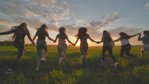 Eleven cheerful girls run to meet the sun across the field in summer holding hands.