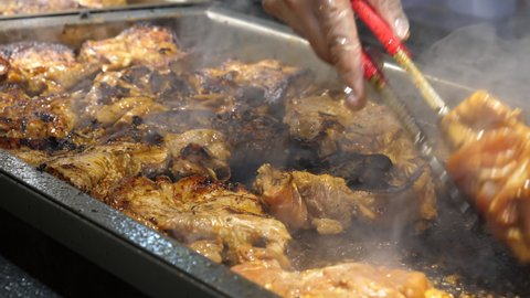 Close up view of chef's hand moving steaming cooking pieces of Jamaican jerk chicken with kitchen tongs at take away or takeout stall.