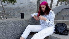 Beautiful red-haired student girl with smartphone sitting on the steps in the city happy to receive good news on social networks.