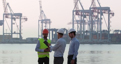 A seaport worker talks to men in white helmets. Engineers approach the worker and communicate against the backdrop of the seaport and the sea.