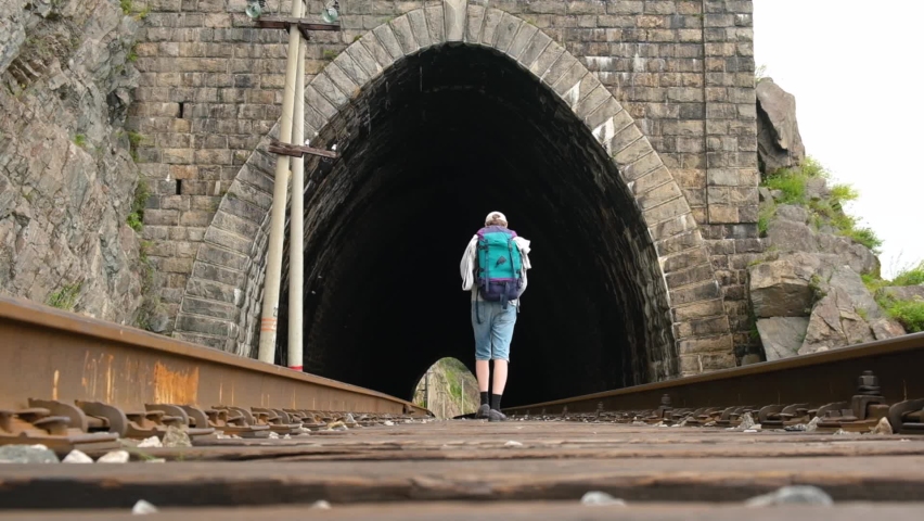 A male tourist with backpack walks along the rails of railway laid through tunnel. Slow motion. Local travel, exploring new places and hiking on weekends. Active recreation, the concept of tourism.