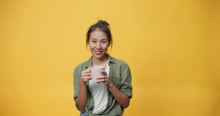 Happy beauty Asia youth lady hold cup of tea or coffee stand over isolate yellow empty space background looking at camera feel fresh and dreamy relax give sign presenting advertisement smile excited. Royalty-Free Stock Footage #1078849922