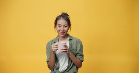 Happy beauty Asia youth lady hold cup of tea or coffee stand over isolate yellow empty space background looking at camera feel fresh and dreamy relax give sign presenting advertisement smile excited.