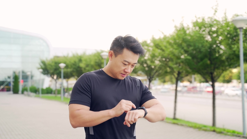 Sport asian man looking smart watch at modern track. Man runner using smartwatch bracelet. Portrait of fitness man checking result. Male in sportswear jogging in the street urban background city park Royalty-Free Stock Footage #1078850030