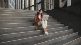 Caucasian young attractive business lady sitting on stairs outdoors near office center working on laptop and have a video call. Woman talking on video conference online outdoors.