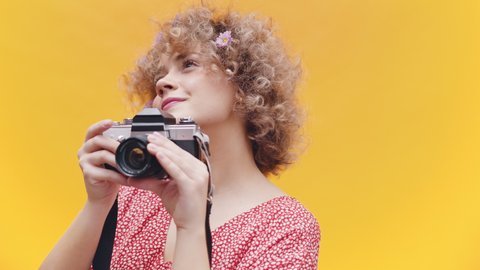 Girl holding a vintage camera in a joyful mood. A camera hanging around her neck. Bright yellow background. Dressed in a casual pink shirt wearing flowers in her hair. Photography concept.