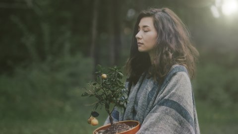 Portrait young hipster female wrapped in a grey plaid in forest early warm morning. Girl vegetarian holding a pot with fresh green plant and look sadly at camera. Nature protection greenpeace concept