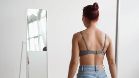 Slim Happy Woman in Tank Top and Jeans Runs Up Mirror Look at Herself. Attractive Girl Looks in Mirror Choosing What Wear at Home on White Background. Smiles and Enjoys Himself Enjoying Good Morning.