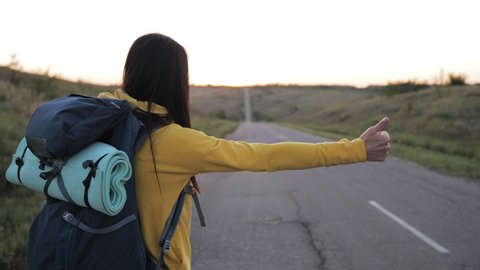 The concept of traveling and hitchhiking. A young female traveler hitchhiking and stopping car with thumbs up on road. A female hitchhiker by the road during vacation trip. Summer vacation adventure.