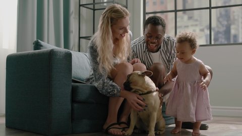 Different races family couple with toddler daughter pets small dog on sofa with cushions against big window in room slow motion