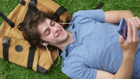 Top view of young man in earphones having video call on cellphone lying on grass outdoors. Student with backpack talk online on video chat relaxing on lawn in park