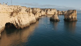 Aerial shot of group of people practicing sup boarding at Ponta de Piedade, Portugal, Lagos, Europe. Overhead view of sporty women and men paddleboarding in ocean at cliffy coastline, 4k footage