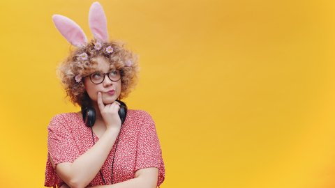 Cute girl wearing bunny ears hairband and spectacles. Headphones around her neck. Thinking something looking away from the camera