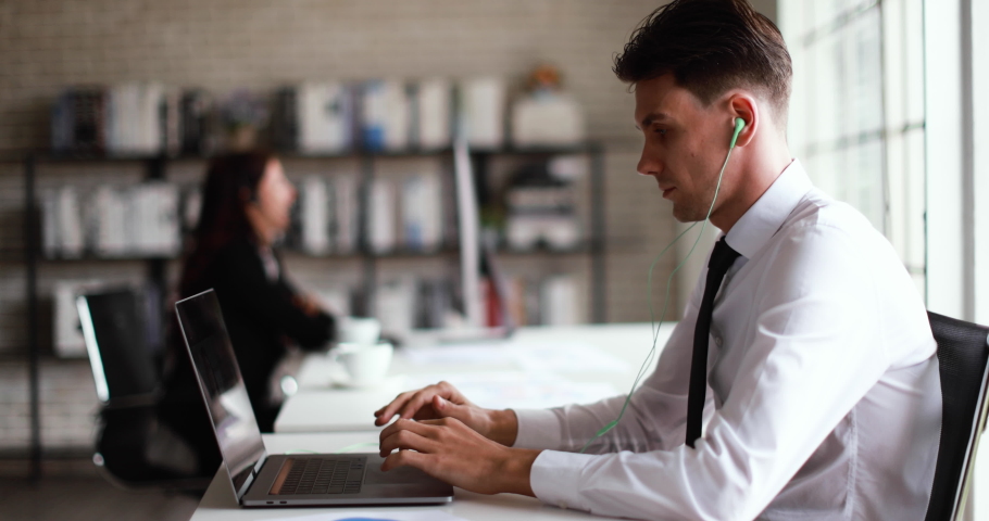 Smart young businessman sitting at desk and talking with team in online conference in office with another businesswoman speaking with other internet meeting session in blur background.