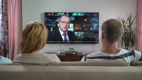 Family Couple Watching TV News Sitting on Couch in Living Room Together. TV Presenter Telling Breaking News about Politics, Elections and Voting Violations