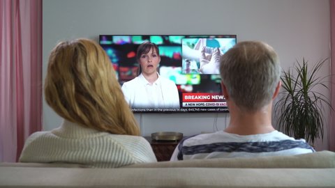 Family Couple Watching TV News Sitting on Couch in Living Room Together. TV Presenter Telling Breaking News about Coronavirus, Lockdown and Vaccinations. Rear View with Dolly-in