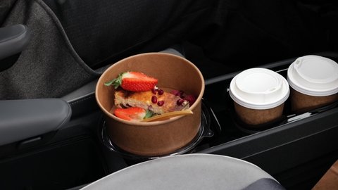 take away coffee cup and cottage cheese casserole dessert with strawberries, pomegranate seeds on panel of automobile in car holder. Multitasking driving concept. Coffee to go, take away healthy food