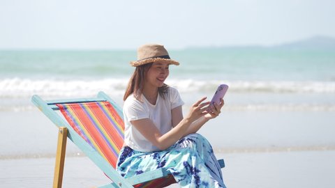Asian woman resting on sunbed on the beach. Happy girl sitting on beach chair by the sea using smartphone taking selfie or video call. Female enjoy beach outdoor lifestyle activity on summer vacation