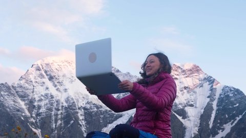 Young happy freelance woman in the mountains of outdoor at sunset calls up by video with her friends via a laptop and shows them all the beauty around