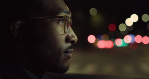 Cinematic close up shot of handsome afro american successful elegantly dressed businessman with glasses is driving modern luxury car in center of the city by night.