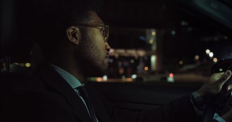Cinematic shot of young handsome afro american successful elegantly dressed businessman with glasses is driving modern luxury car in center of the city by night.
