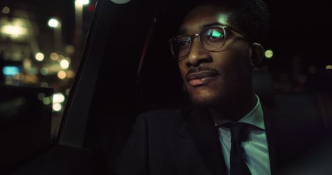 Cinematic shot of handsome afro american successful elegantly dressed businessman with glasses is looking outside window during traveling in luxury modern car with driver in center of the city by nigh