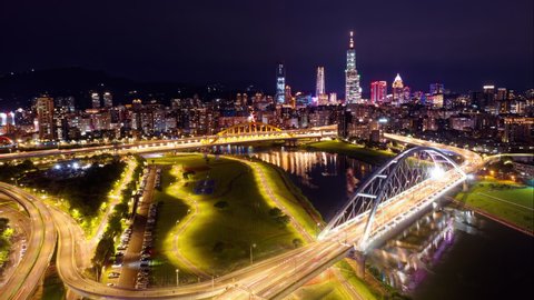Night skyline of Taipei City in aerial hyperlapse, viewed from above a riverside park, with highway bridges over Keelung River and 101 Tower among the dazzling lights in Xinyi Financial District