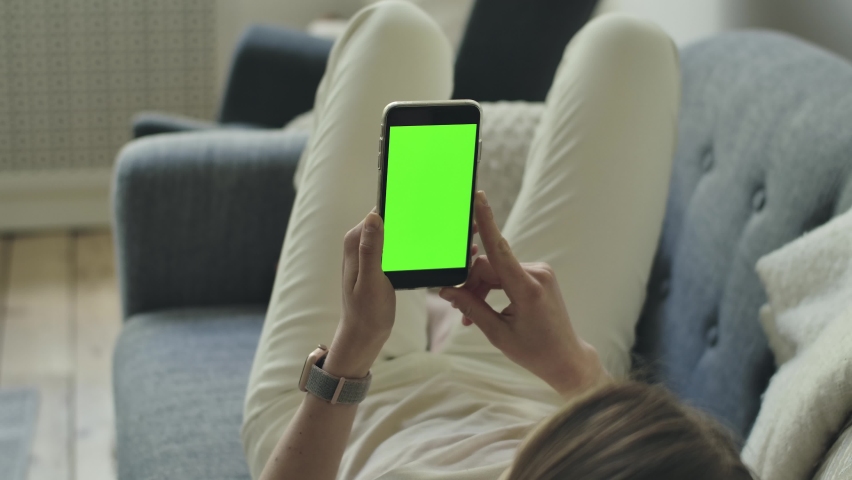 Female person watches smartphone in hands. Empty chromakey display with mock up space for advertisement logos, texts. Woman lies on sofa, looking at mobile phone with green screen for ads, adverts. Royalty-Free Stock Footage #1078905842