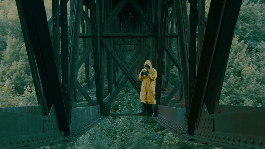 A man in a yellow protective suit, gas mask, black leather gloves stands under an iron bridge and sets fire to an orange smoke bomb.
