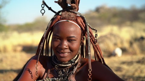 Beautiful Himba woman looking at camera and smiling, wearing traditional jewellery and headdress in her village near Kamanjab in northern Namibia, Africa.
