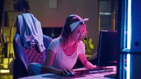 Young woman wearing headphones playing computer game, winner. Female gamer in glasses winning hard match, looking at computer monitor, using computer mouse and keyboard. Cybersport, gaming club