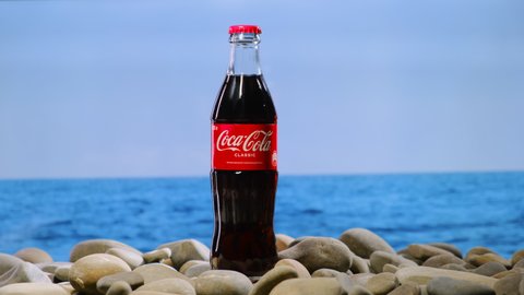 Moscow, Russia - CIRCA 2021: man open glass bottle of Coca Cola with silver shiny opener. concept of summer vacation on stone beach with sea or ocean in background. Packshot of soda.