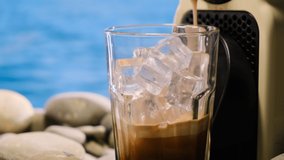 Close up iced coffee early morning at sea, making latte on vacation by ocean. Beige capsule coffee machine makes coffee in transparent mug or cup with ice. relaxing on beach, in background blue water. - Powered by Shutterstock - Get 15% off with code: PIKWIZARD15