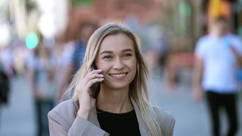 Portrait of trendy blonde European woman speaking on smartphone. Stylish portrait of smiling city businesswoman chatting on mobile phone. Crowded cellphone talk. Smart people hold phones in hands.
