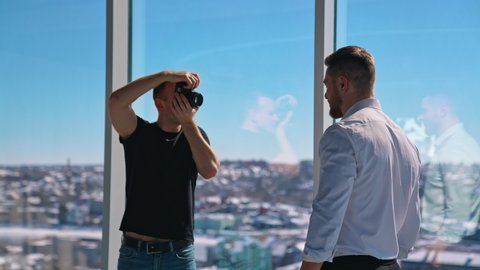 Young man working with camera in office. Businessman posing on camera. Professional photographer taking photos of a male model. Photo session near window.