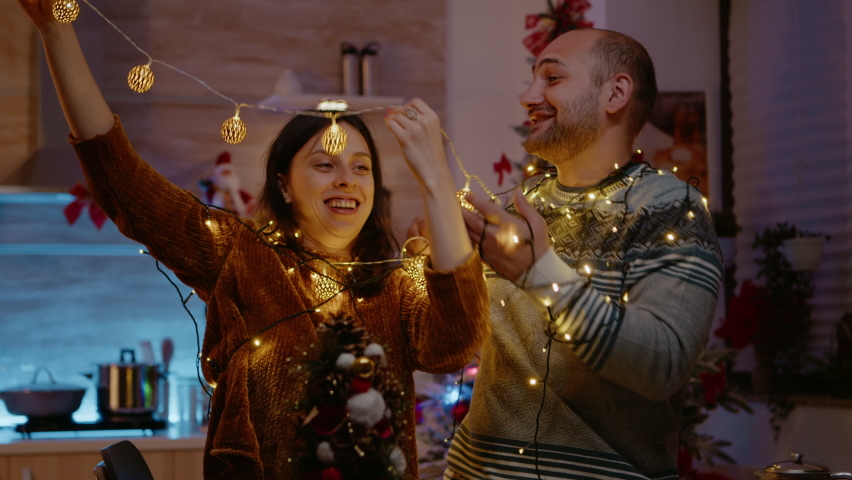 Festive couple knotted in string of christmas lights. Man and woman laughing while trying to untangle garland with twinkle lights and illuminated bulbs for holiday celebration.
