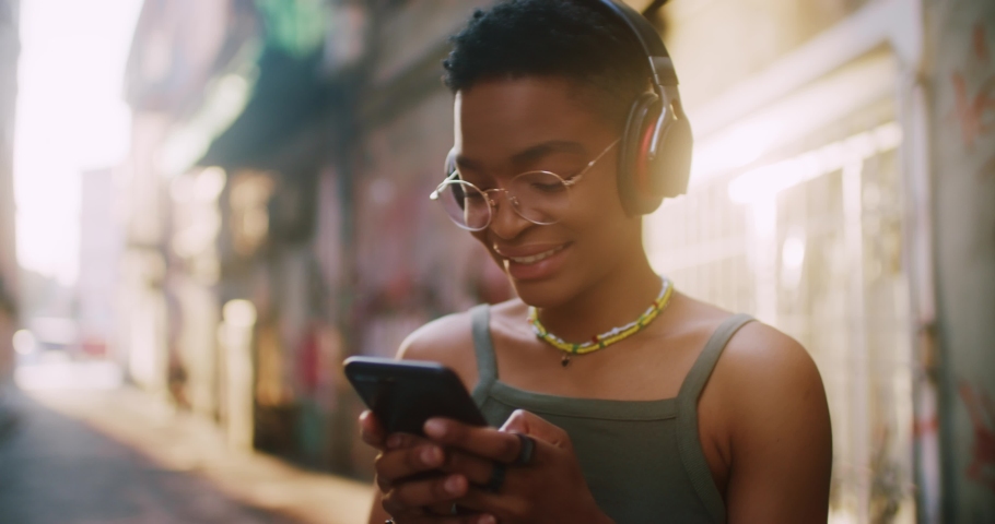 Portrait young african american girl enjoying listening to music stand on street at sunlight use phone. Look at camera. Woman, joyful, positive, enjoyment. Slow motion Royalty-Free Stock Footage #1078945796