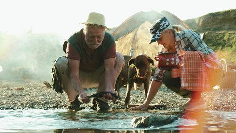 Old man cleaning fresh fish in a stream on a beach, prepares food in nature. A happy retired couple and dog is camping by the river and preparing fish for dinner. Concept of survival in nature