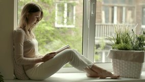 Girl with novel book in hands reads while sitting by opened window on sill. Female reader in cozy home at reading hobby. Rest with literature stress relief. Woman sits on windowsill with drama story. - Powered by Shutterstock - Get 15% off with code: PIKWIZARD15