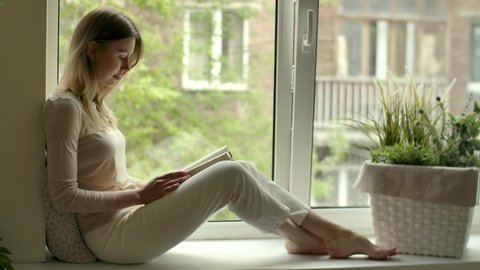Girl with novel book in hands reads while sitting by opened window on sill. Female reader in cozy home at reading hobby. Rest with literature stress relief. Woman sits on windowsill with drama story.