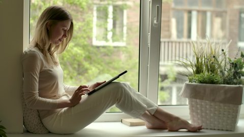 Girl with computer tablet in hands draws while sitting by opened window on sill. Female artist in cozy home at sketching hobby. Rest with drawing stress relief. Woman sits in windowsill with e-tablet.